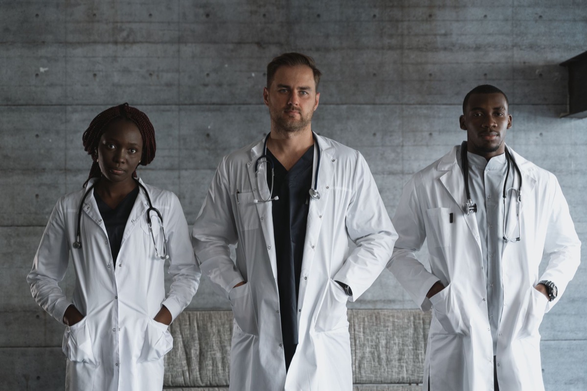 Medical team in white coats standing in a sterile examination room with a white examination table and a digital monitor displaying 'Best Specialists'